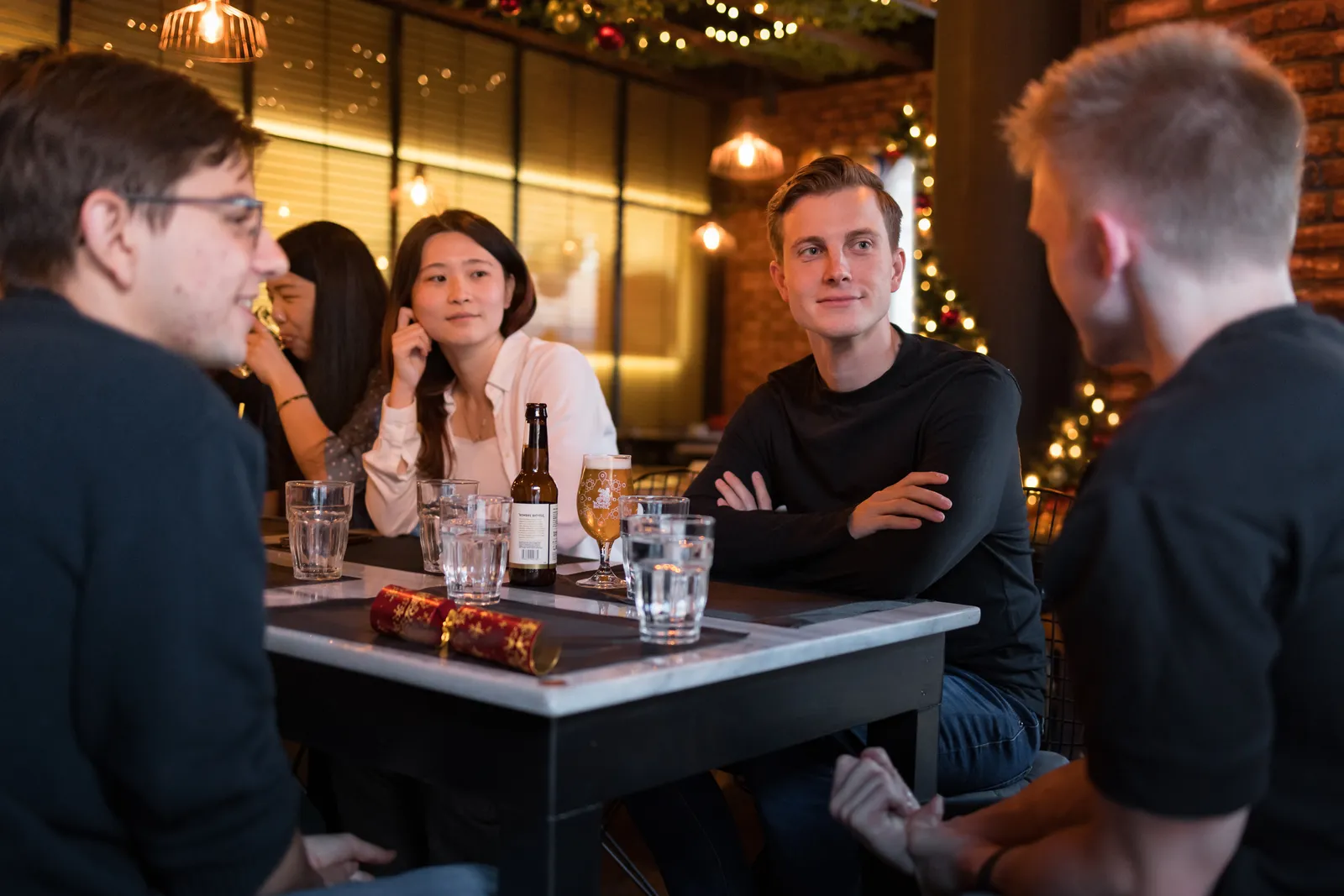 Friends round a table at Padharo with drinks and a Christmas cracker, fairy lights and a lit tree behind them
