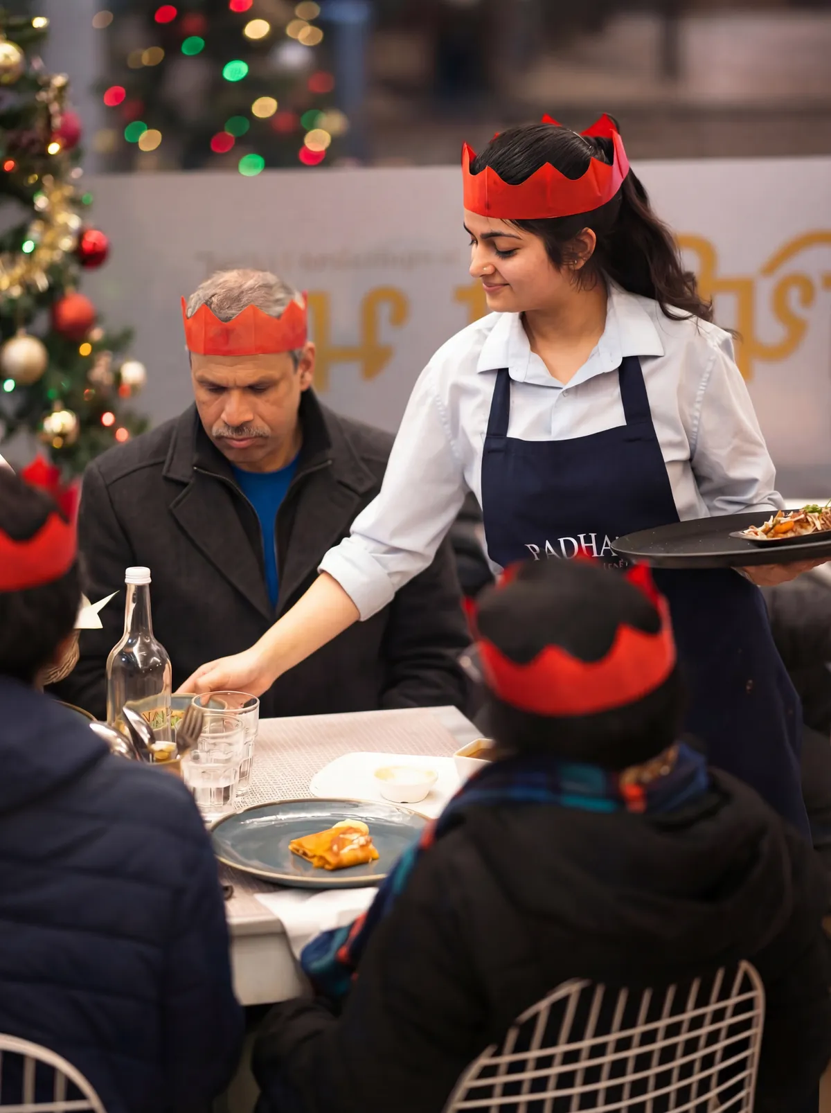 A member of our team in a paper crown serving a dosa to a family table at Christmas, with the tree lit behind them